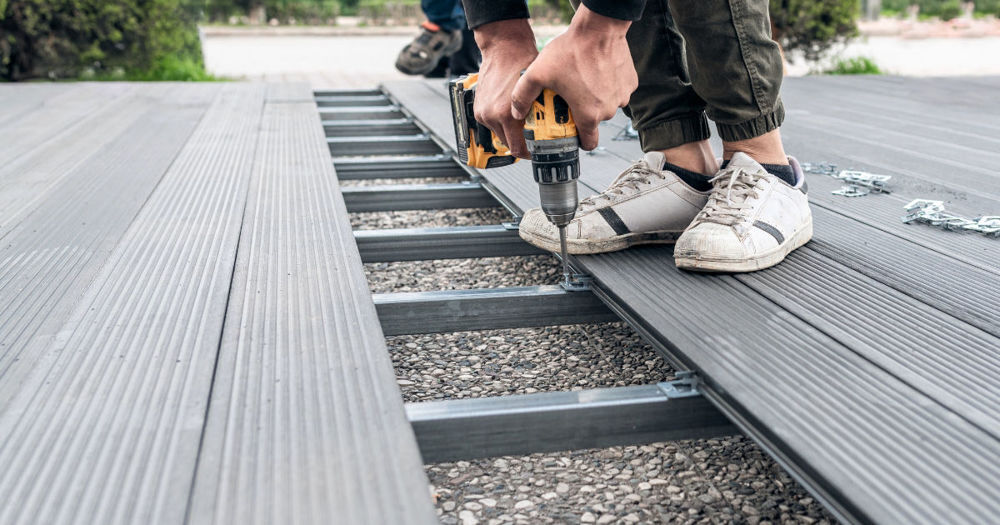 The image shows a close-up view of the construction of an outdoor deck with grey composite decking material. A person is in the process of installing the deck, using a drill to secure the decking boards to the metal frame underneath.